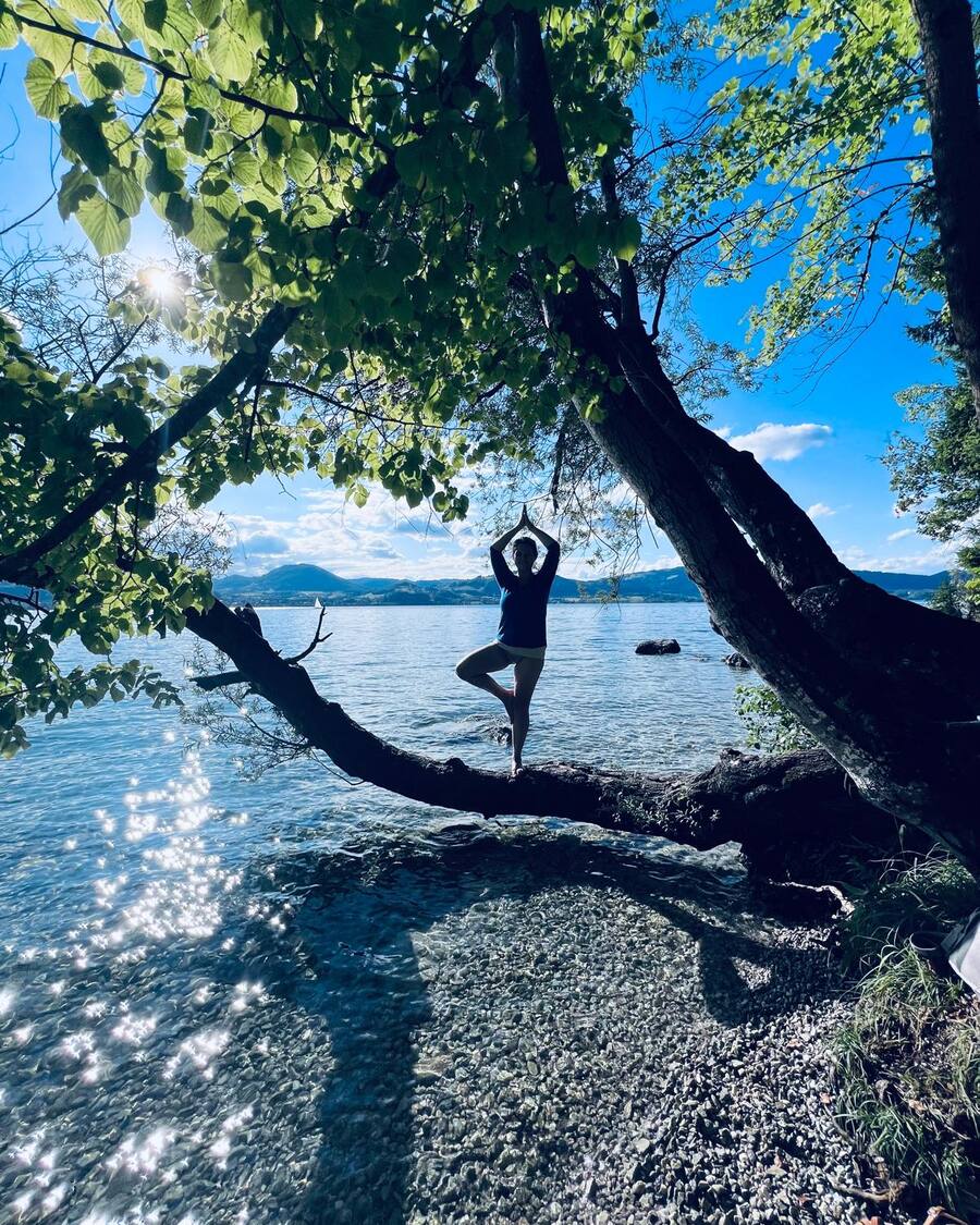 Dora steht im Baum auf einem Ast über dem See, die Hände über dem Kopf, dahinter Berge