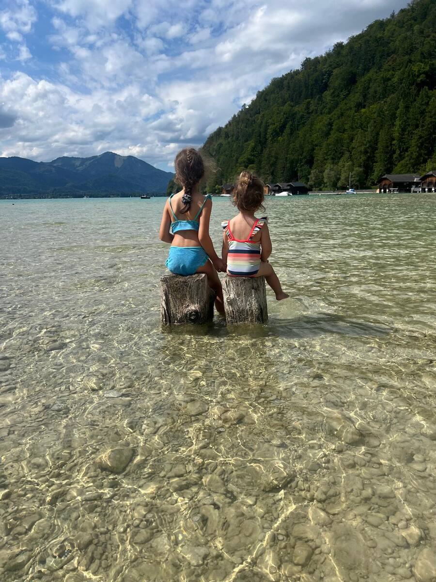 Zwei Kinder sitzen von hinten auf Holzpfählen im flachen See, dahinter Berge und Wald