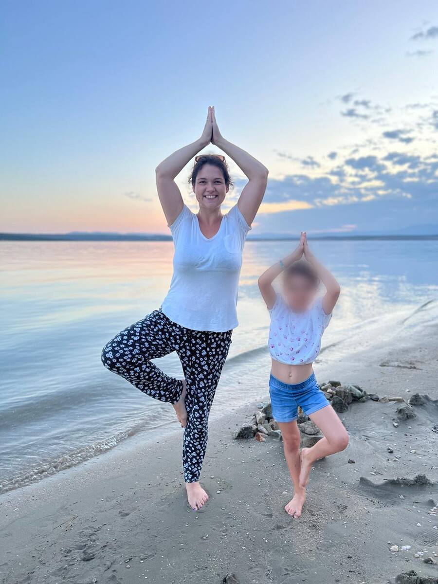 Dora und ein Kind stehen barfuß am Strand in der Baumhaltung, die Hände über dem Kopf, dahinter das Meer im Abendlicht